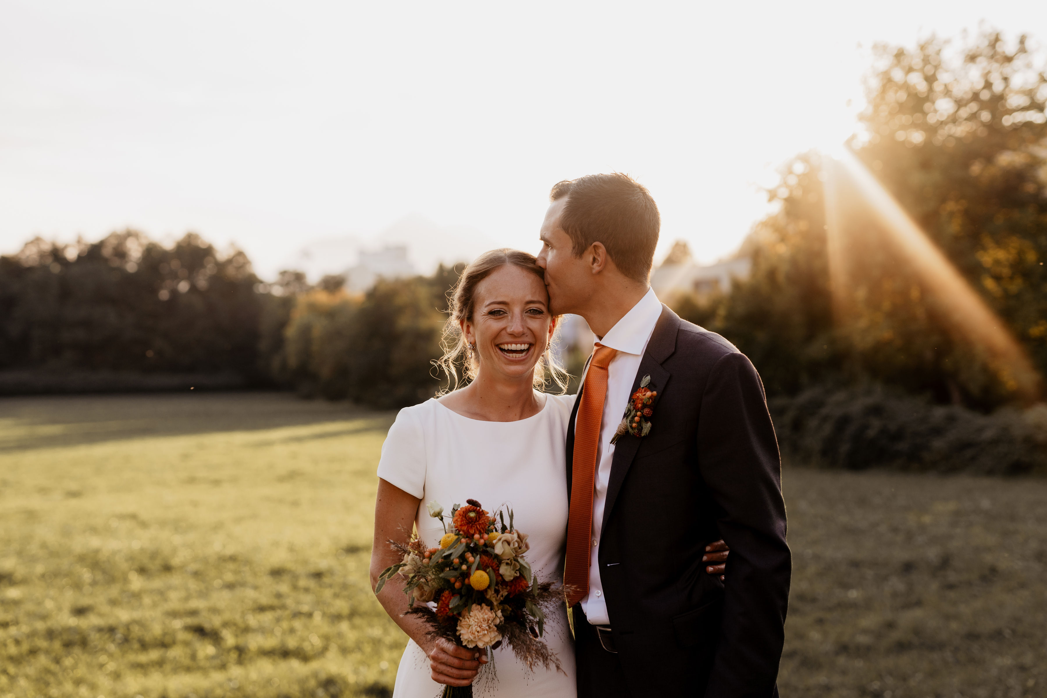 Liebevoller Augenblick: Bräutigam küsst Braut auf die Schläfe, während sie lacht und einen Brautstrauß hält.